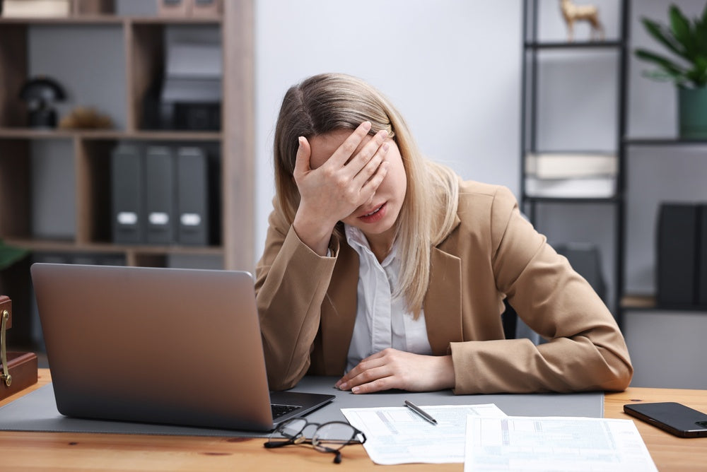 Woman at a wooden office desk in the afternoon, eyes closed, resting her head on her hand